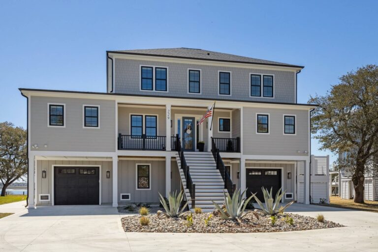 Three-story coastal custom home with dual garages, a central staircase, and a neutral exterior palette accented by black trim and drought-tolerant landscaping.