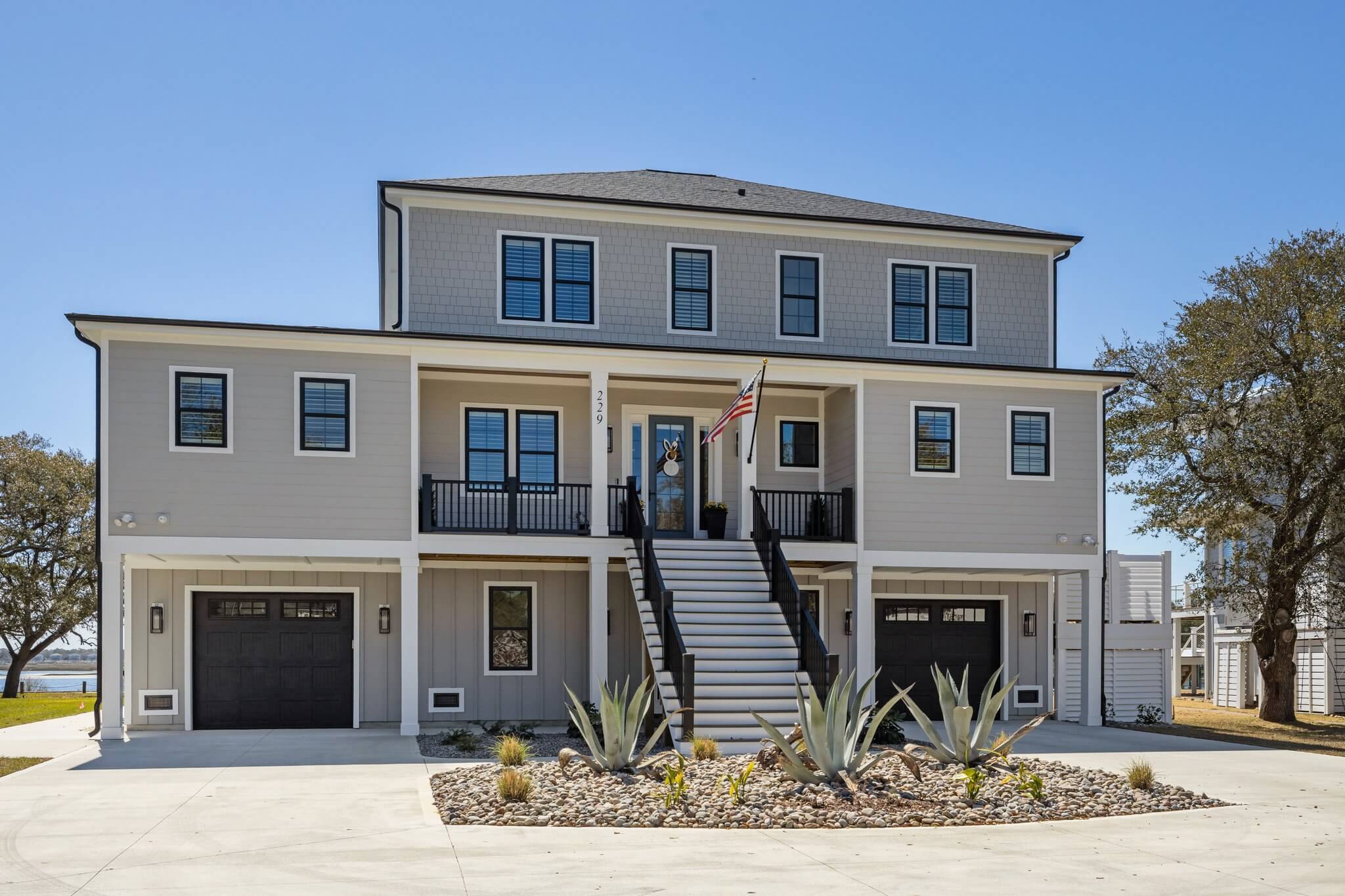 Three-story coastal custom home with dual garages, a central staircase, and a neutral exterior palette accented by black trim and drought-tolerant landscaping.